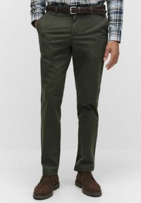 Man i olivgröna chinos med ett flätat brun bälte, en rutiga skjorta instoppad, och mörkbruna mockaskor, står med avslappnade händer.