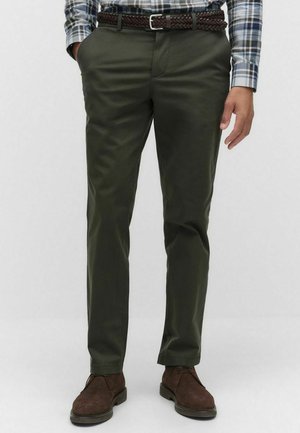 Man in olijfgroene chino's met een gevlochten bruine riem, een ingezoomd geruite shirt en donkerbruine suède schoenen, staande met ontspannen handen.