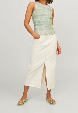 Femme portant un haut vert sans manches, une jupe midi blanche fendue devant, et des tongs vertes, debout avec une main dans la poche.