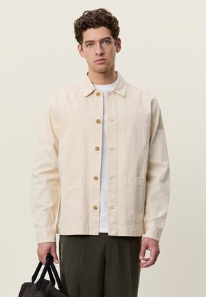 Homme aux cheveux bouclés portant une veste beige boutonnée sur une chemise blanche, tenant la poignée d'un sac noir dans la main gauche sur un fond uni.