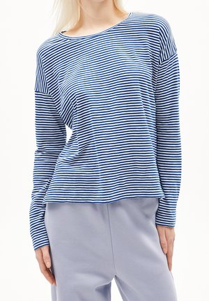Femme portant une chemise à manches longues rayée bleu et blanc associée à un pantalon gris clair sur fond blanc.