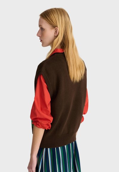 Jeune femme aux longs cheveux blonds portant un pull sans manches marron, une chemise rouge à manches longues et une jupe rayée bleue, regardant vers la gauche.