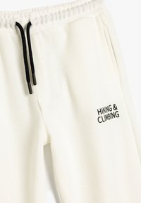 Pantalones de chándal blancos con textura acanalada, cinturilla elástica y cordones negros. Presenta un texto bordado en negro que dice "HIKING & CLIMBING."