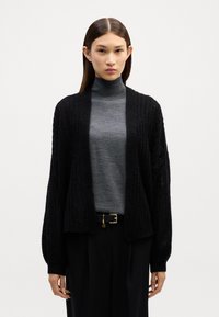 Črna pletena kardigan s odprtim sprednjim delom, rebrasto teksturo in širokimi rokavi, oblečena čez sivo turtleneck majico, v kombinaciji s črnimi hlačami.