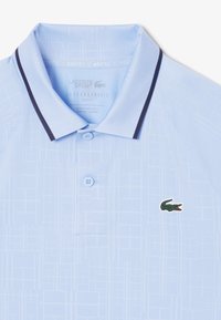 Ljusblå kortärmad pikétröja med ett texturerat mönster, svarta kragekanter och en grön logotyp på vänster bröst. Har två knappar.