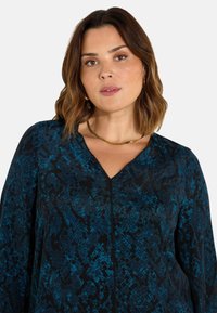 Blůza s výstřihem do V v černé a tyrkysové barvě, vyrobená z texturované tkaniny s geometrickým vzorem; má dlouhé rukávy a jemný lesk.