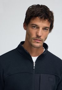 Svart fleecejacka med dragkedja, i strukturerad tyg, med krage och en logotyp på bröstet. Har en slät dragkedjestängning och neutral bakgrund.