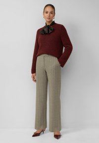 Femme debout dans un pull en tricot marron, blouse noire avec nœud, pantalon à carreaux beige et talons aiguilles bordeaux sur fond gris uni.