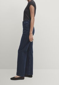 Jeans a gamba larga blu scuro in denim, con vita alta e tasche anteriori. Abbinati a una t-shirt nera aderente e scarpe basse.