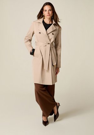 Femme portant un trench-coat beige ceinturé sur un top noir et un pantalon large marron, posant avec une main dans la poche et des escarpins noirs pointus.