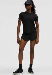 Vrouw in een zwart sportoutfit met een pet, zonnebril, shorts en sneakers, staande tegen een effen grijze achtergrond.