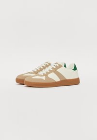 Tan- og hvide sneakers med en tekstureret gummisål, der har grønne detaljer og tre diagonale striber på siderne. Snøredesign.