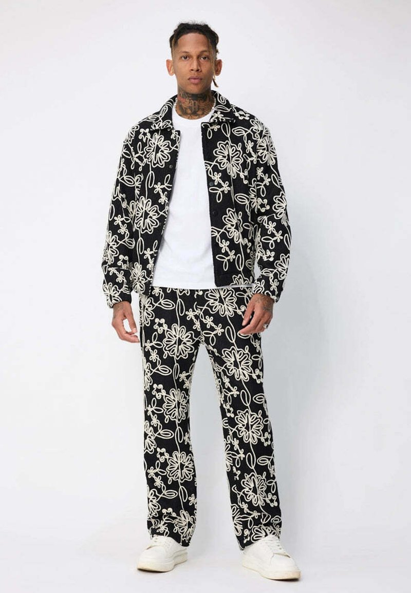 Homme portant une veste et un pantalon noirs assortis avec de grands motifs floraux blancs, une chemise blanche et des baskets blanches sur un fond uni.
