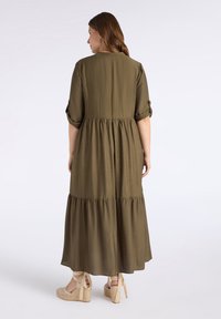 Olivgrünes Maxikleid aus weichem Stoff, mit einem gestuften Design, rundem Ausschnitt und hochgerollten Ärmeln. Getragen mit Wedge-Sandalen.