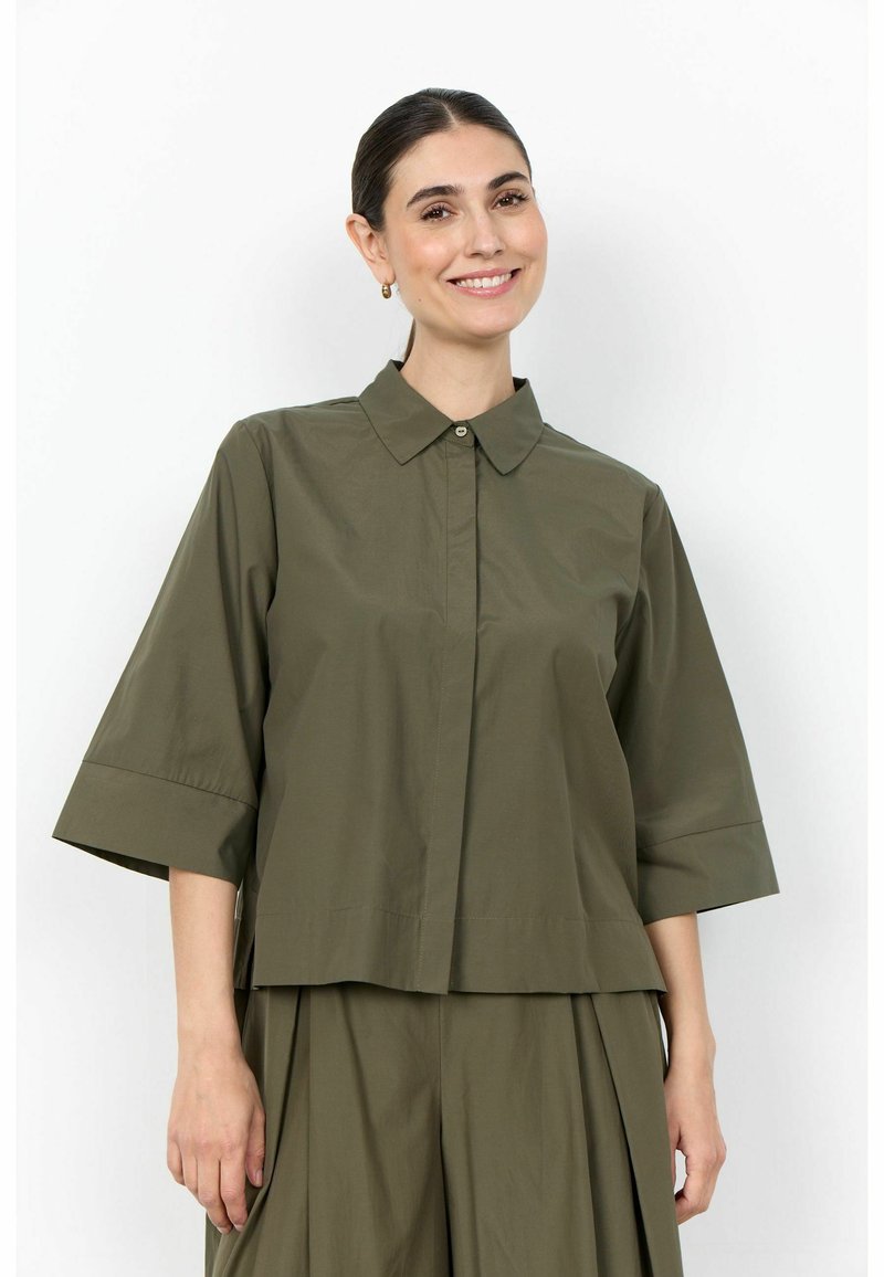 Femme souriante aux cheveux foncés attachés en un chignon bas porte une chemise ample vert olive à manches mi-longues et une jupe plissée assortie.