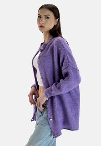 Fioletowy sweter na drutach z zaokrąglonym dekoltem, zapinany na guziki z przodu, o luźnym kroju i ściągaczowych mankietach, noszony na białej bluzce i niebieskich dżinsach.