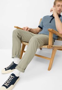 Korte mouw, marineblauw-wit gestreepte polo, beige broek, marineblauwe sneakers met crèmekleurige zolen, zittend op een licht houten stoel tegen een witte achtergrond.