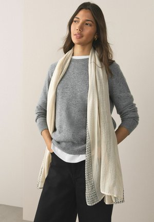 Junge Frau trägt einen grauen Pullover, eine weiße Bluse, schwarze Hose und einen hellbeigen Schal mit dezenten Mustern, steht mit den Händen in den Taschen.