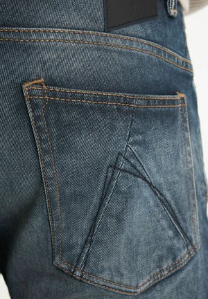 Hintertasche von blauen Denim-Jeans mit orangefarbenen Nähten und einem markanten DoppelChevron-Schwarzstich-Design.