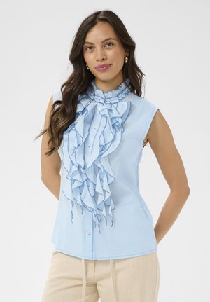 Femme aux longs cheveux noirs portant un chemisier sans manches bleu clair à volant sur le devant et un pantalon beige, debout devant un fond uni.