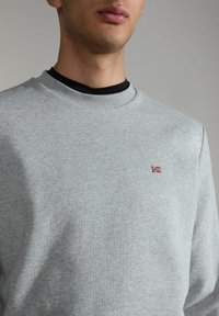 Sudadera gris claro hecha de un tejido suave. Presenta un cuello de canalé y un pequeño emblema de bandera roja y azul en el pecho. Textura lisa.