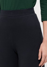 Pantalons noirs côtelés avec une large ceinture élastique. Le tissu est texturé, offrant extensibilité et confort. Main positionnée sur le côté.