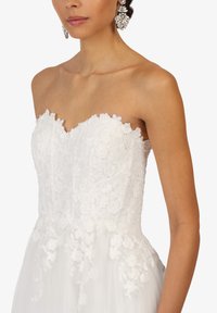 Robe blanche sans bretelles avec des détails en dentelle florale, jupe en tulle et décolleté festonné. Modèle portant des boucles d'oreilles chandeliers.
