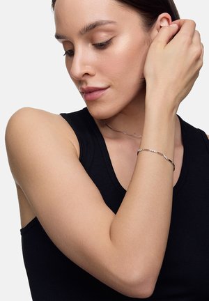 Srebrna bransoletka z okrągłymi, przezroczystymi akcentami; elegancki, cienki design noszony na odkrytym nadgarstku. Modelka ma na sobie czarny top na ramiączkach, prezentując biżuterię.