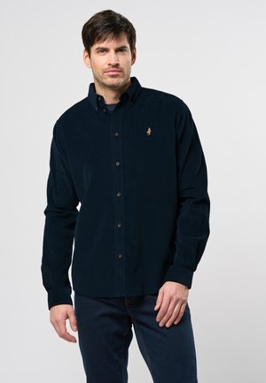 Man in een donkerblauwe corduroy overhemd met knopen en een klein geborduurd logo op de borst, gecombineerd met donkere jeans, staand tegen een witte achtergrond.
