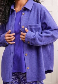 Fialová fleecová košile s knoflíkovou légou, jednou náprsní kapsou a dřevěnými knoflíky. Nositelná přes odpovídající fialový top a vzorované kalhoty.