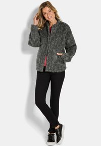 Vrouw in een fuzzy grijze jack met knopen, rode top, zwarte skinny broek en zwarte sneakers staat met één hand in de zak en glimlacht.