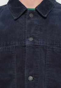 Μπουφάν από μπλέ ναυτικό κορδούρα με κλασικό γιακά και τέσσερα κουμπώματα. Υφασμα με υφή και κάθετες ραβδώσεις.