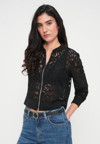 Μαύρο μπουφάν bomber με περίτεχνο σχέδιο δαντέλας με λουλουδάτα μοτίβα, εμπρόσθιο κλείσιμο με φερμουάρ και ριμπ μανσέτες και στρίφωμα. Συνδυασμένο με μπλε τζιν.