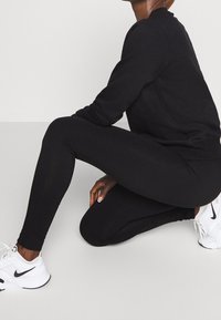 Sudadera negra y mallas hechas de tela elástica. Zapatillas deportivas blancas con detalles en negro. La persona está arrodillada sobre un fondo neutro.