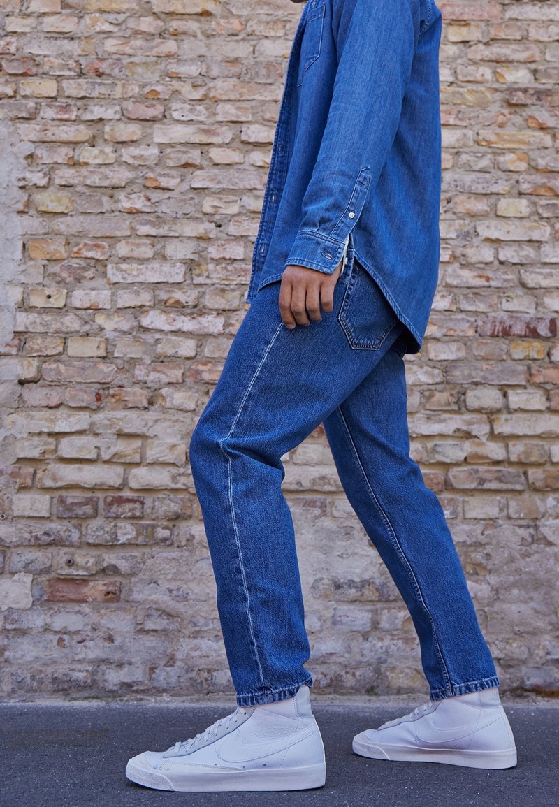 Džínová košile a džíny v odstínech modré, s volným střihem. Bílé vysoké tenisky dotvářejí outfit. Pozadí tvoří kamenná zeď.