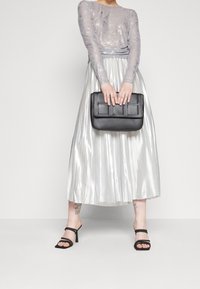 Silberne plissierte Rock mit einem eng anliegenden, glitzernden Langarmoberteil. Hält eine schwarze rechteckige Clutch-Tasche mit strukturierter Oberfläche. Schwarze Absatzsandalen.