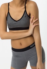 Kobieta w szarym biustonoszu sportowym Nike i pasujących szortach, z założonymi rękami, stojąca na tle białego tła.