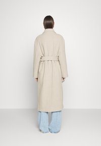 Beige wollen jas met een ceintuur in de taille, lange mouwen en een brede kraag. Draagt over lichtblauwe uitlopende spijkerbroek, gezien van achteren.