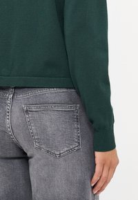 Grønn strikket genser med ribbestrikkede mansjetter, parret med grå denimjeans som har baklommer og en falmet tekstur.