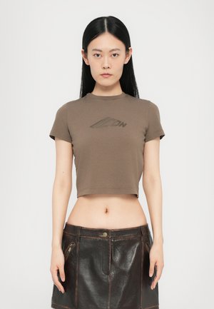 Brun cropped t-shirt med korte ærmer og rund halsudskæring, der har et subtilt logo print over brystet; parret med en mørk lædernederdel.