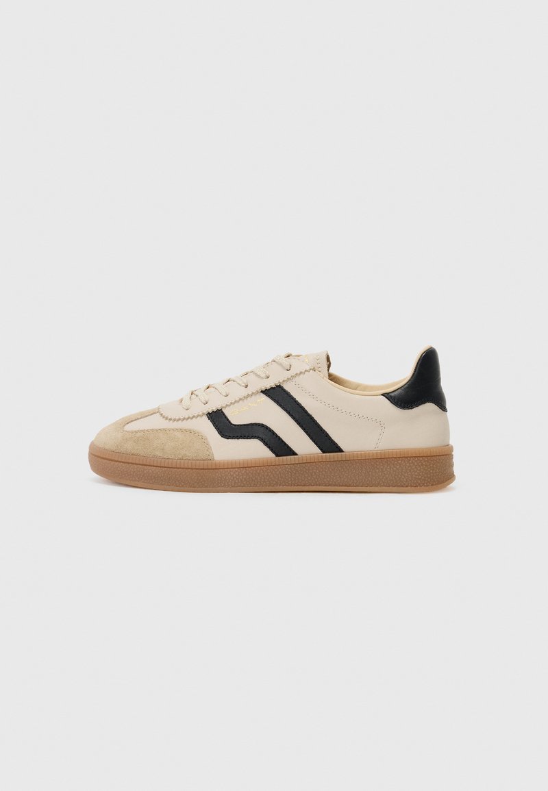 Beige sneakers with black stripes, suede toe cap, and a gum rubber sole. The design features a smooth finish and a lace-up front.