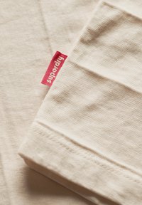 Camiseta de algodón color crema con textura acanalada, que presenta una etiqueta de tela roja con "superdry" impreso en ella. Forma rectangular simple.
