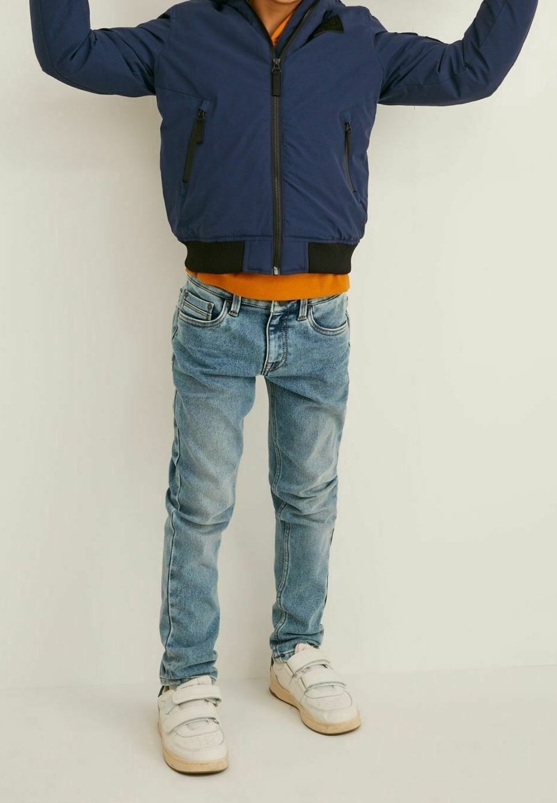 Enfant portant une veste bleue, une chemise orange, un jean bleu clair et des baskets blanches, les bras levés devant un fond uni.
