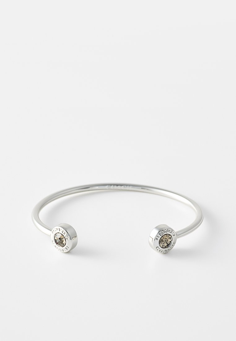 Brazalete de puño de plata con dos extremos acentuados con cristales. Presenta una forma redonda y suave con un acabado pulido. Diseño minimalista con detalle de logo.