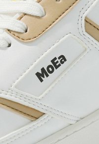 Biele tenisky s béžovými akcentmi, vrch z texturovanej kože a biele šnúrky. Obsahujú čierne logo "MoEa" na boku a podrobné šitie.