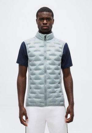 Uomo che indossa un gilet trapuntato grigio chiaro sopra una camicia blu navy a maniche corte e pantaloni bianchi, in piedi davanti a uno sfondo semplice.