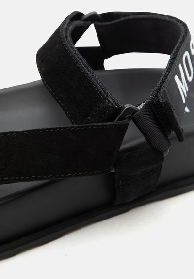 Μαύρο σανδάλι με ρυθμιζόμενους ιμάντες Velcro και λευκό κείμενο στον ιμάντα του αστραγάλου, με μαξιλαρωμένη σόλα και ανθεκτική ραφή.