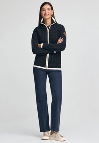 Kobieta stoi ze skrzyżowanymi ramionami, ubrana w granatowy sweter na zamek z warkoczowym splotem, ciemne jeansy i jasnobeżowe trampki na jednolitym tle.