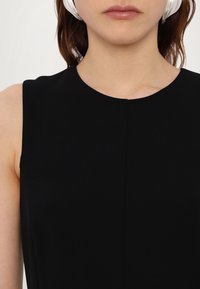 Femme portant un haut noir sans manches à encolure ronde, avec des écouteurs blancs sur les oreilles.