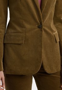 Rudas kordorojinis švarkas su vertikaliu raukšlėtumu, vieno mygtuko užsegimu ir dviem priekinių kišenių. Pritaikytas fasonas su lygiu pamušalu.